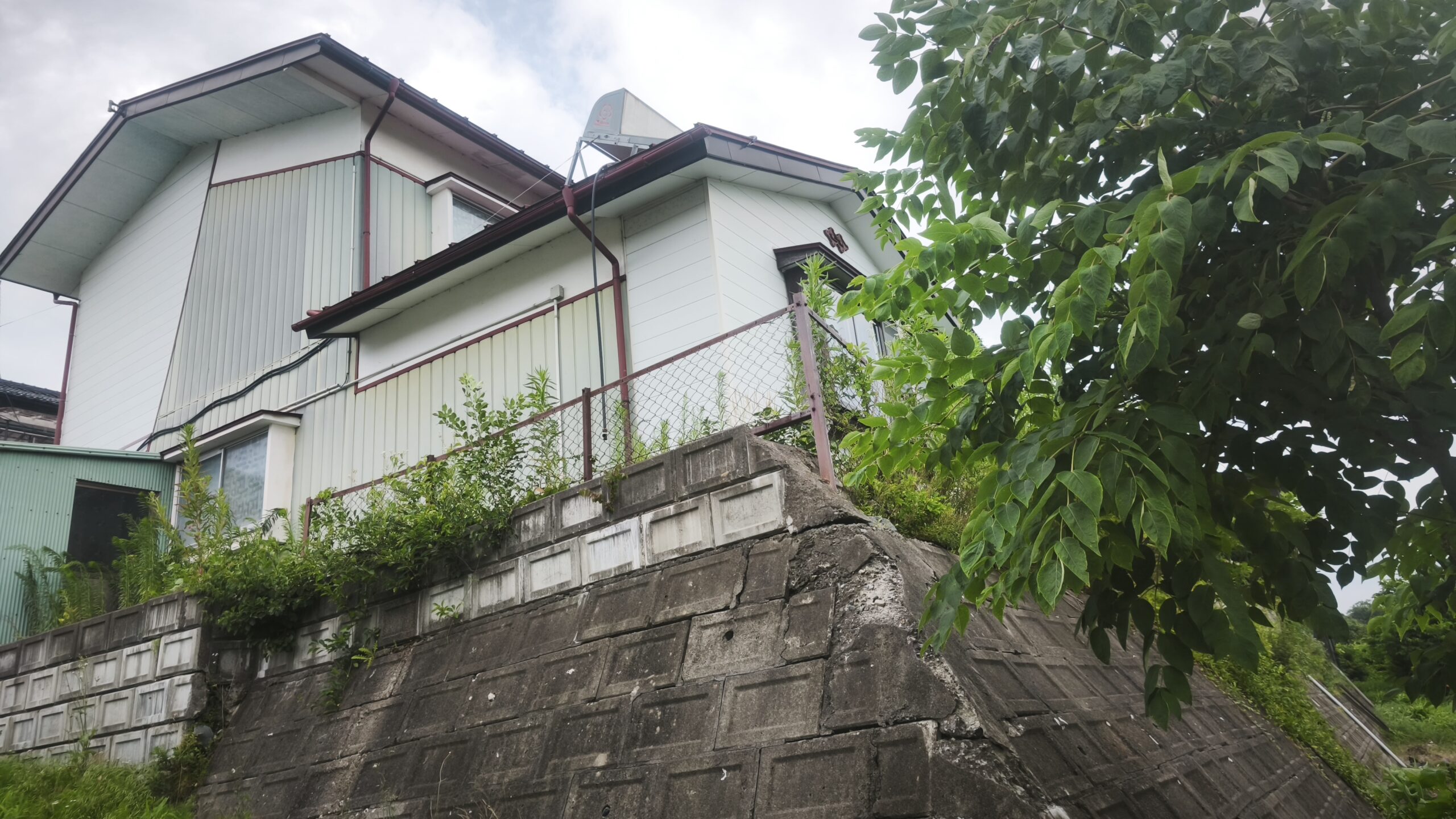 岩手県遠野市の実家の外観。石垣の上に建つ木造2階建てで、庭木や雑草が伸びている